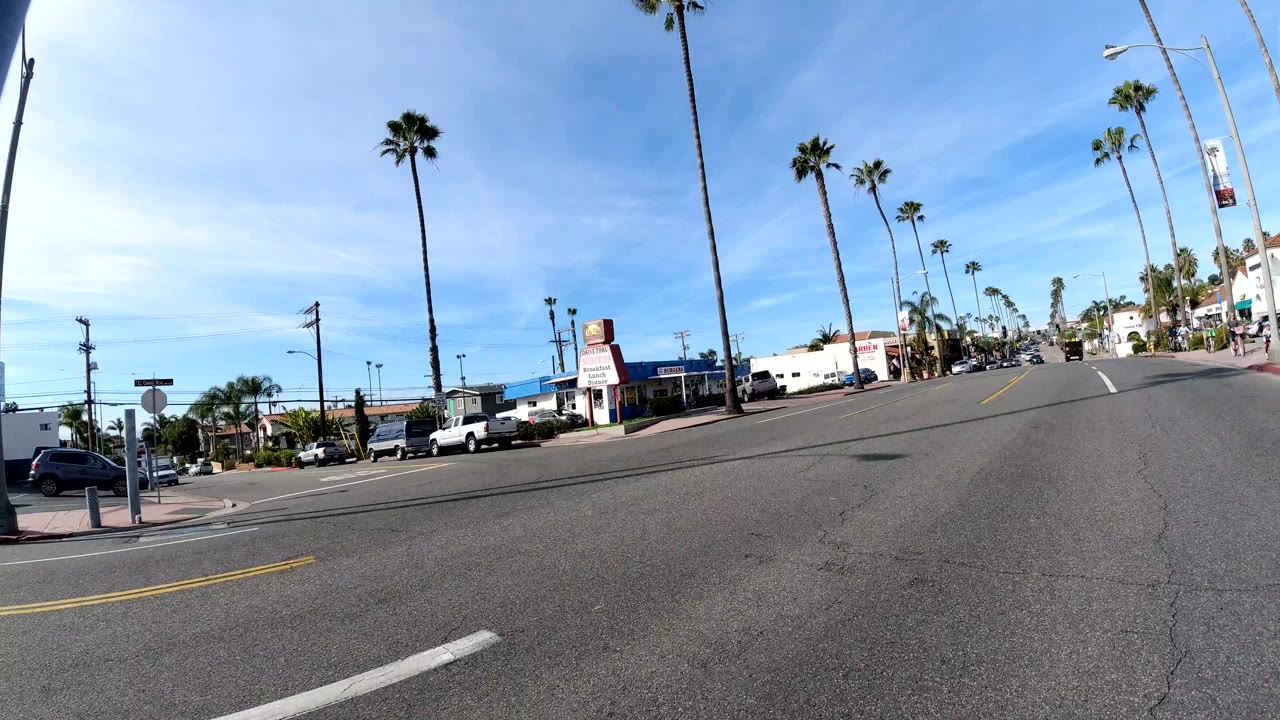 Roadside view in San Clemente looking along a broad street with palm trees, a roadside business sign, parked vehicles, traffic in the distance, and tiny pedestrians near the far sidewalk.