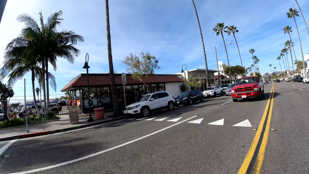 Palm-lined street with restaurant-style storefront, parked cars, and red pickup truck.