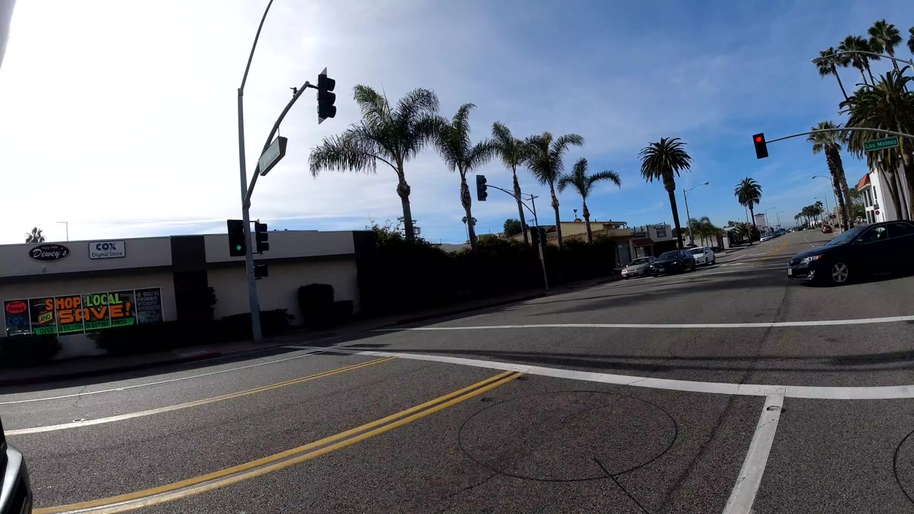 Onboard camera at 0:00 on a San Clemente avenue: a dark gray pickup truck fills the right foreground, palm trees and traffic signals ahead, commercial signage on the far left, blue sky with scattered clouds; no people are clearly visible