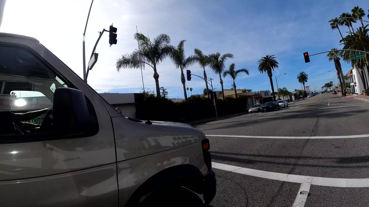 Close dashcam view with the gray pickup truck side mirror prominent in the right foreground and palm fronds overhead