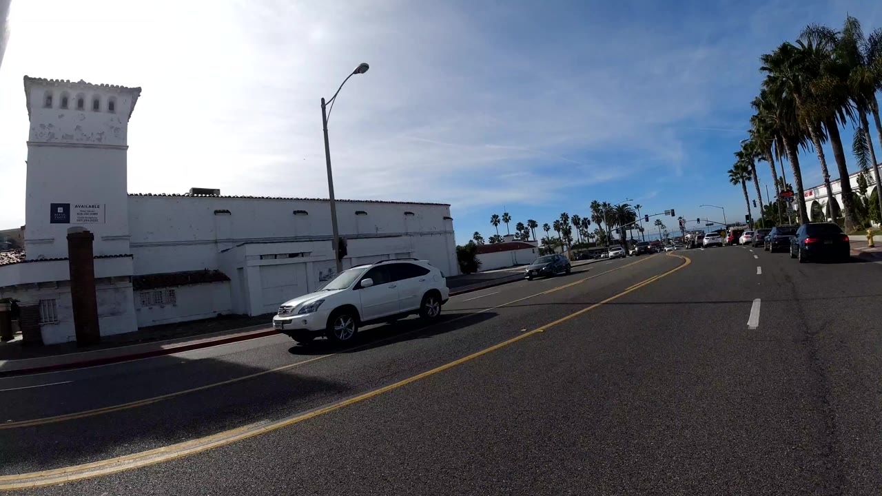At 0:02 the pickup clears and the San Clemente avenue opens: a white vehicle ahead in the center lane, a white rectangular building on the right, palm trees and street lamps on both sides of the road, open blue sky; no people are clearly visible