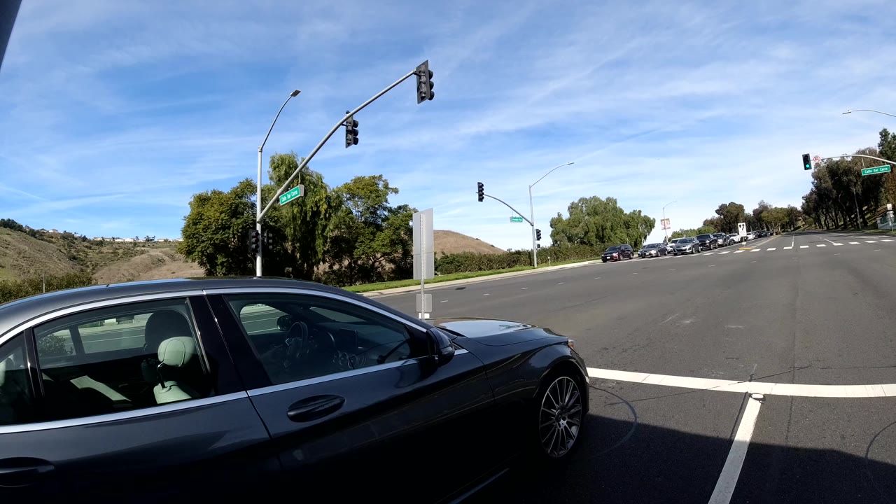 Forward dashcam view in San Clemente, California at the clip's opening showing a dark sedan near a signalized intersection, traffic light arms visible overhead, rolling green hills under a partly cloudy blue sky, no visible people