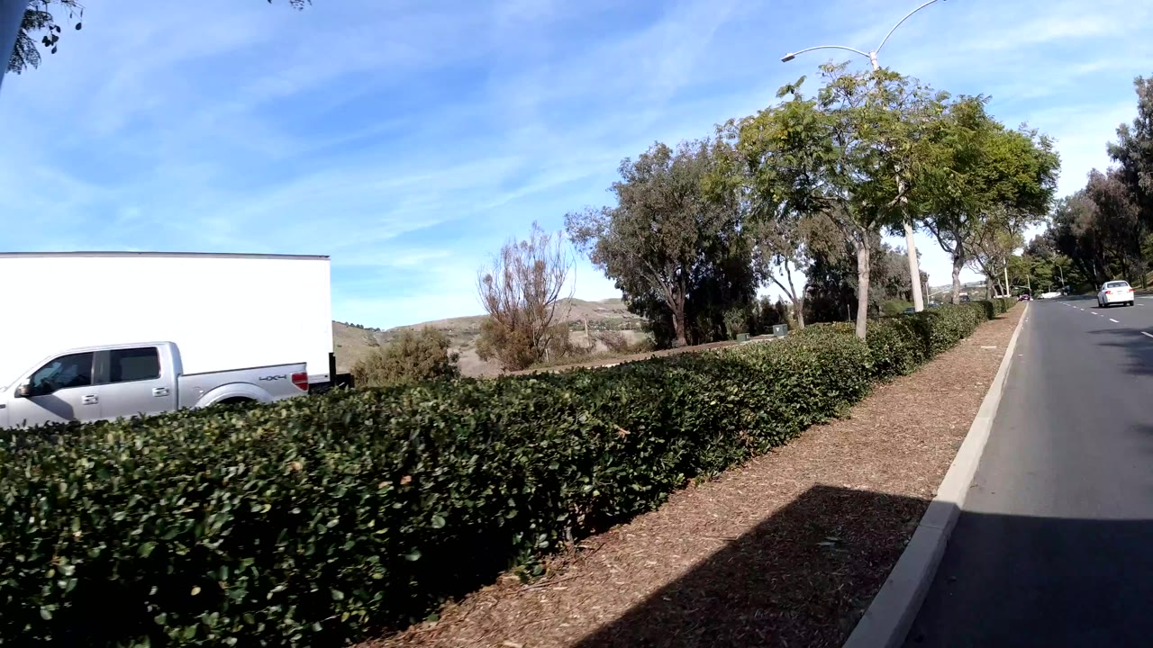 San Clemente street at two seconds with the camera past the intersection; a white pickup or box truck crosses ahead, a dense green hedge lines the right shoulder, rolling tan-and-green hills fill the background, no visible people