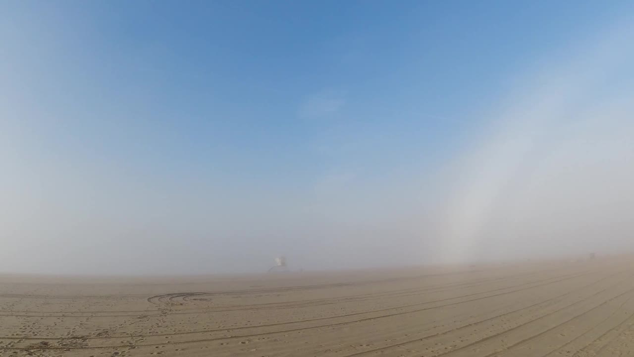 Wide flat sandy terrain with parallel tire tracks in Huntington Beach, California; a white atmospheric arc rises prominently on the left against a hazy blue sky; a small distant structure sits on the flat horizon; no visible people