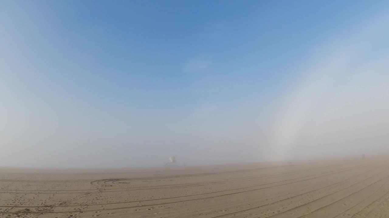Flat sandy plain with tire tracks; white arc moves toward center of hazy blue sky