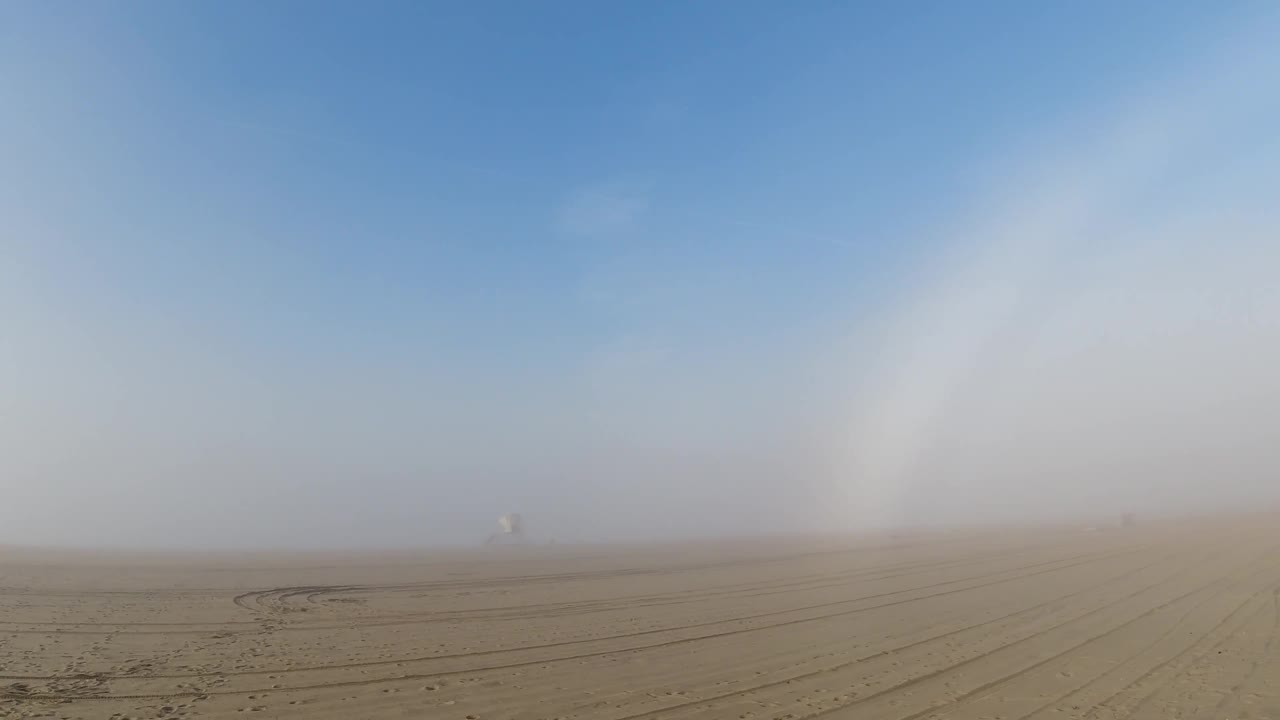The white atmospheric arc has shifted to the upper-right portion of the frame as the pan continues over the flat sandy terrain in Huntington Beach; the sandy foreground shows tire-track furrows; a small distant structure marks the horizon; no visible people