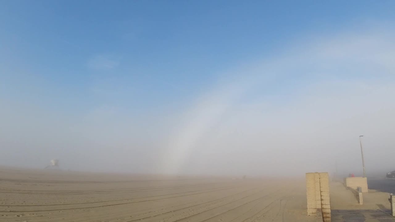 At one second, the fogbow arc holds its position above Huntington Beach's flat sandy terrain; the concrete barrier wall and utility pole remain in the same compositional position and the horizon fog layer appears similarly dense to the opening frame; no people visible