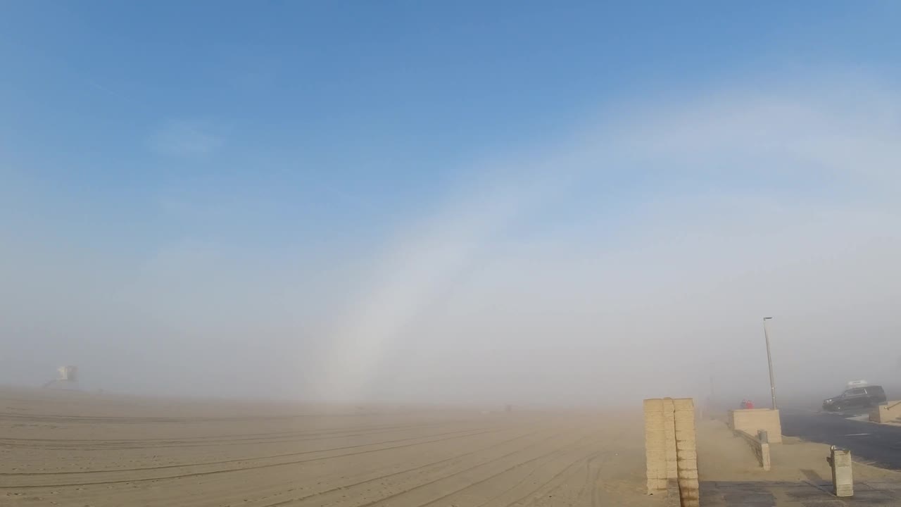 At two seconds above Huntington Beach, the horizon fog layer appears at its densest while the fogbow arc remains prominent overhead; the concrete barrier is still centered in the right foreground and no rightward pan movement toward the road has yet occurred; no people visible