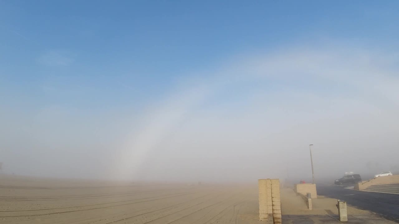 White fogbow over coastal scene, paved road and low distant structure visible behind concrete barrier