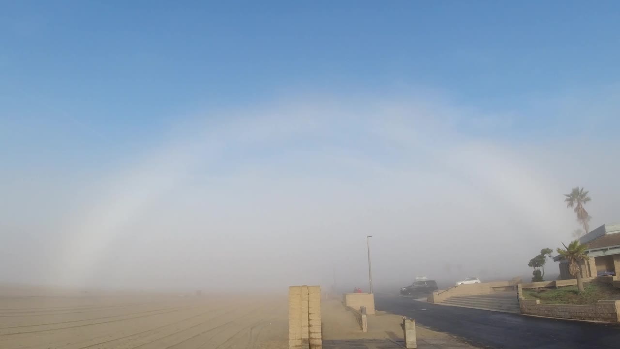 Opening frame showing the full fogbow arc spanning from left to right across the upper sky above a Huntington Beach sandy lot and road, with a concrete post at lower left and a low-rise house with palm trees at right midground; no people visible.