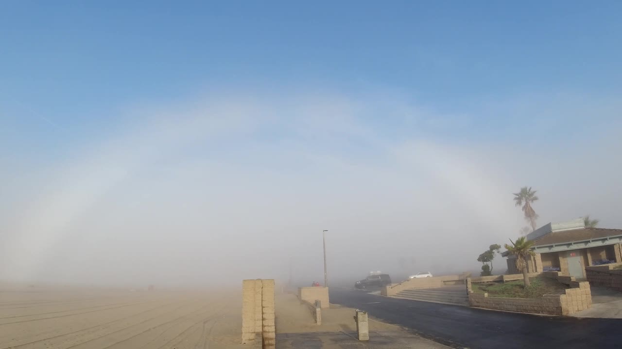 Concrete post dominant at lower left, house and palm trees filling right third of frame, white fogbow arc in upper sky, road and sandy foreground.