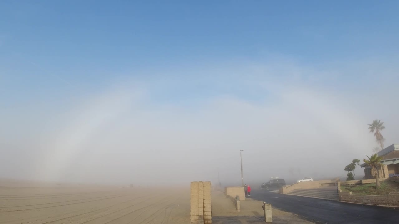 At 0:02 in the Huntington Beach clip, the fogbow arc continues as the primary feature; the residential building and palm trees on the right edge are slightly more defined, while the sandy foreground and hazy sky remain consistent; no visible people.
