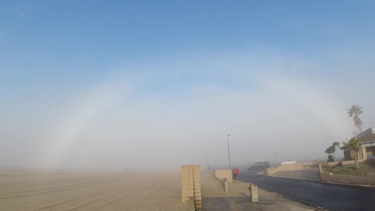 Fogbow arc over sandy coastal road, concrete barrier post prominent in lower foreground