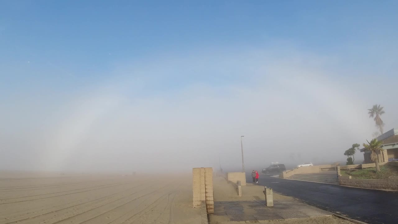 Hazy sunrise view in Huntington Beach with sandy foreground, a dark road at right, low wall pillars, a palm by a house-like structure, and a faint distant red figure or object.