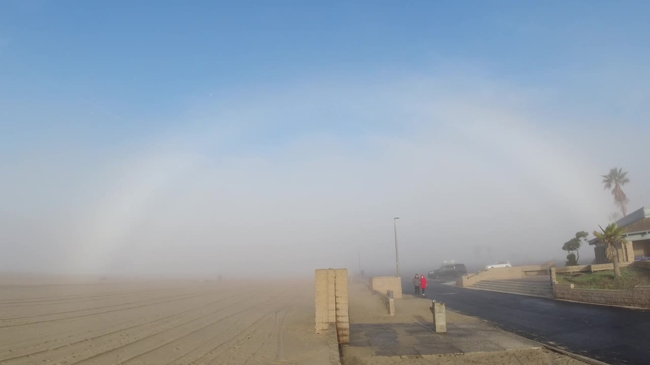 Wide Huntington Beach sunrise frame with sandy terrain stretching left, a paved road and low pillars to the right, a house-like structure, a tall palm, and haze covering the horizon.