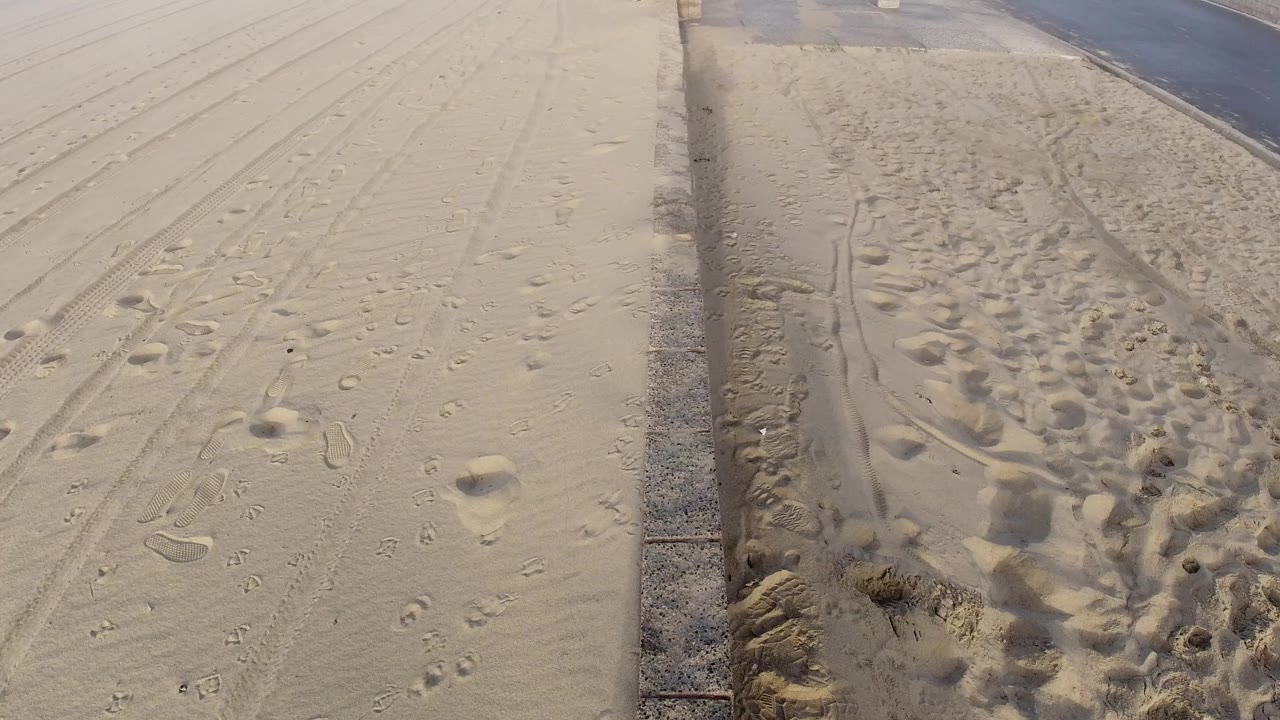 Huntington Beach sand with scattered footprints and track marks beside a paved road, with no visible people in this opening frame.