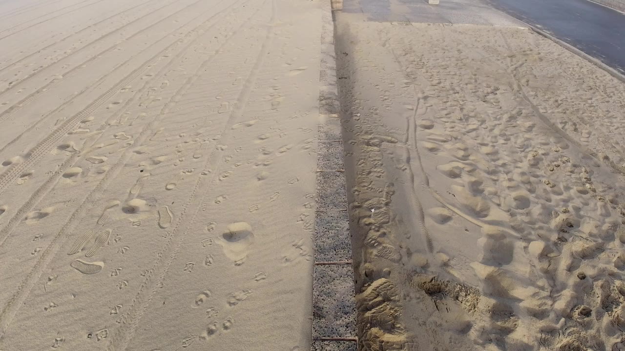 Huntington Beach shoreline-adjacent sand fills the frame with footprints, track lines, a curb, and the road visible at the upper right, with no visible people.