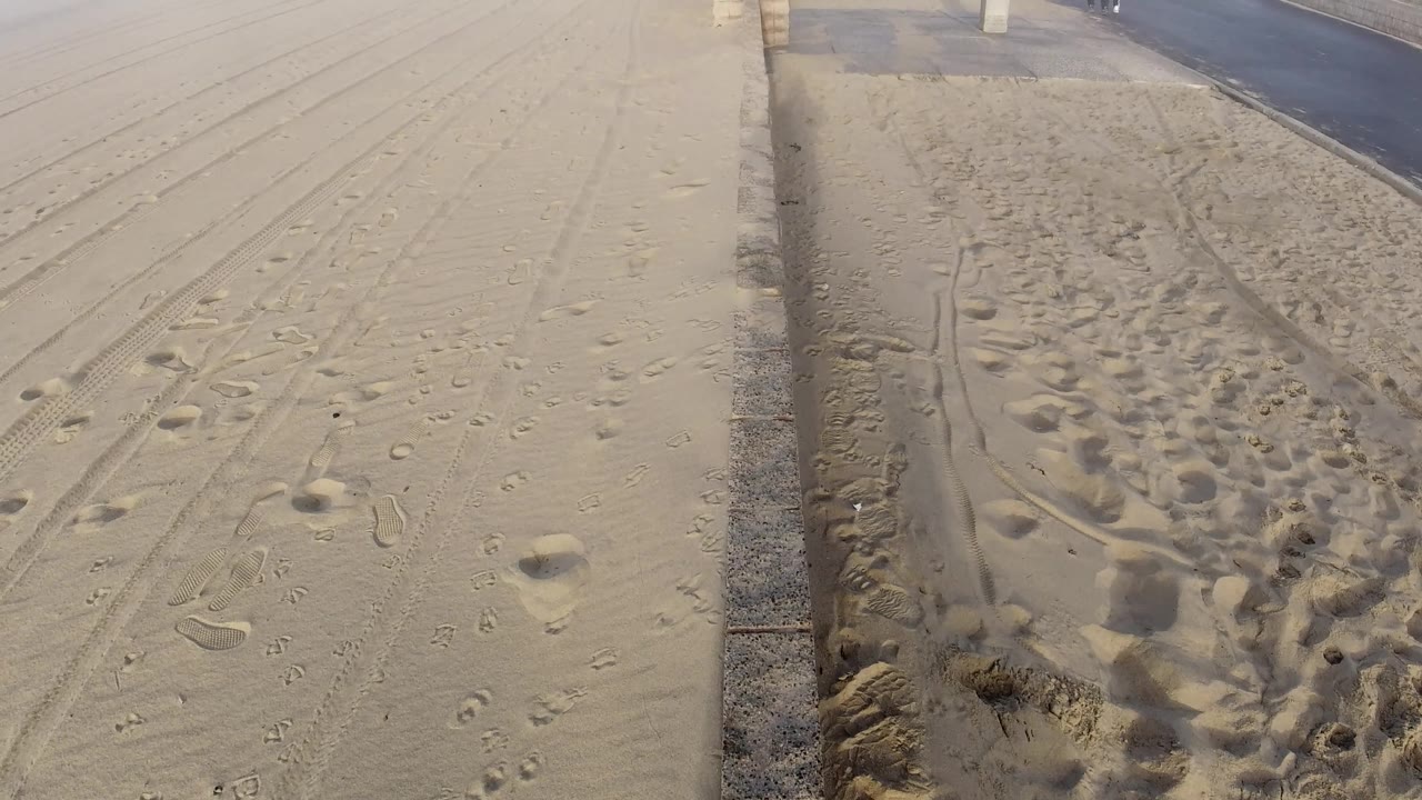 Huntington Beach sand marked by footprints and tracks sits beside a paved road and low concrete posts, with no visible people in the frame.