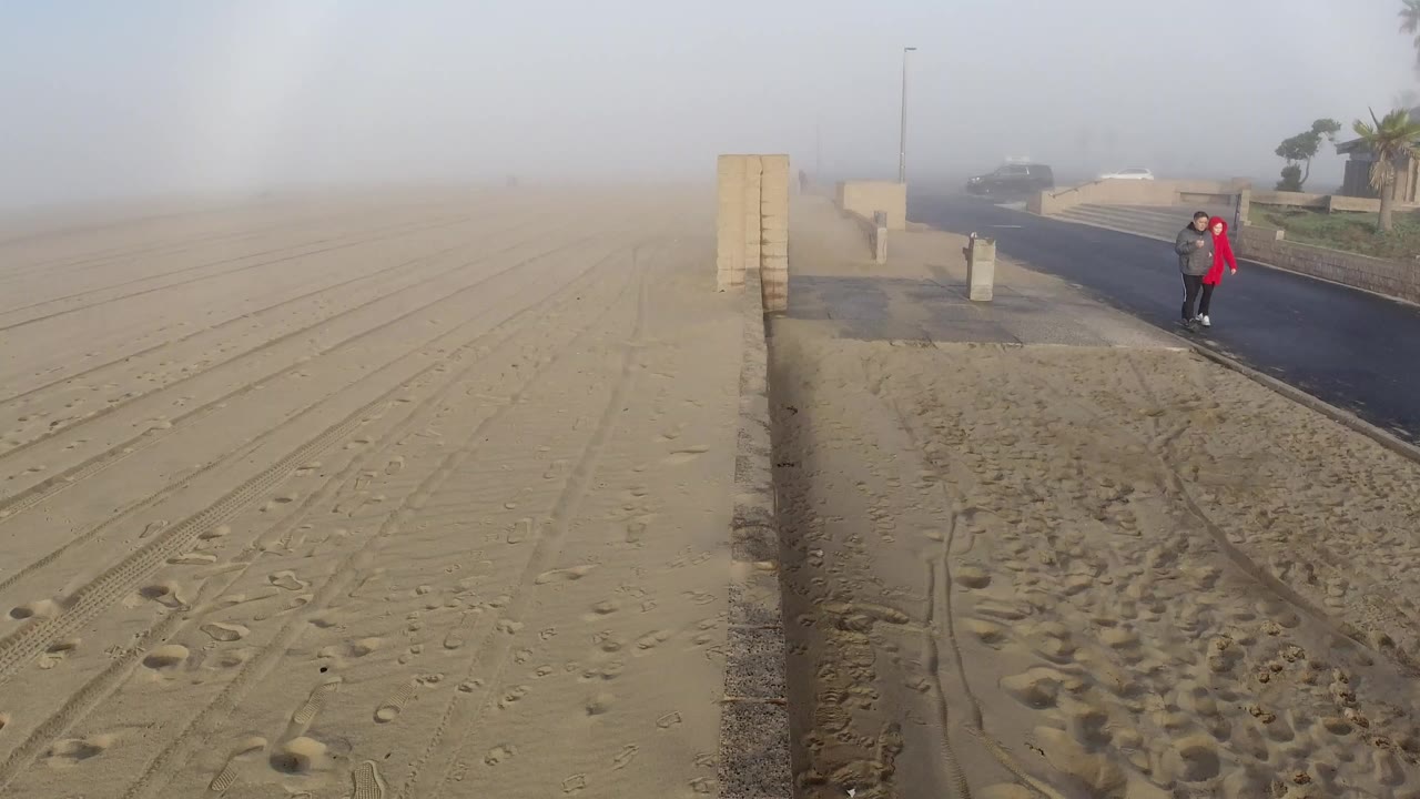 Aerial drone view over Huntington Beach, California, looking down at a sandy beach and coastal road; two people stand on the road, one in a red jacket; the sky shows a diffuse white arc consistent with a sundog; the concrete retaining wall runs diagonally through frame; faces not distinguishable.