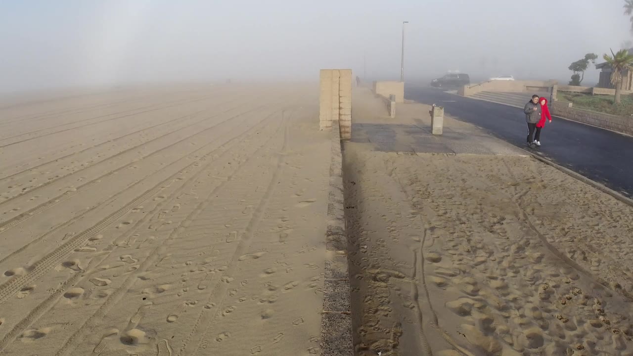 At 1 second the Huntington Beach aerial view is nearly identical to the opener; both figures remain in the same positions on the coastal road, the red jacket visible; the sandy beach fills the lower frame; the atmospheric arc persists in the hazy sky; faces are not distinguishable at this altitude.