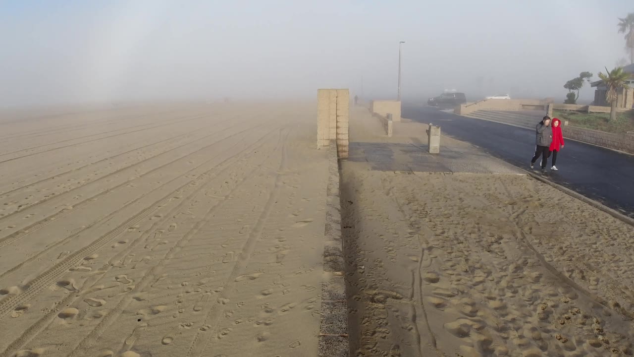 At 2 seconds the two people have shifted slightly in position along the Huntington Beach coastal road; the figure in red is now closer to the retaining wall than in frame 1; the sandy beach and hazy atmospheric arc remain consistent; palm trees visible at upper right; faces not distinguishable.