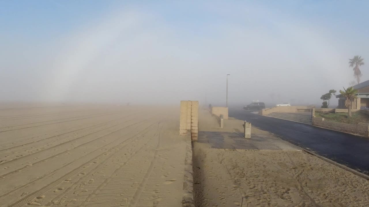 At 0:00 in Huntington Beach, sandy ground with tire tracks fills the left foreground while a paved road, low walls, homes, palms, and hazy gray sky sit to the right, with no visible people.