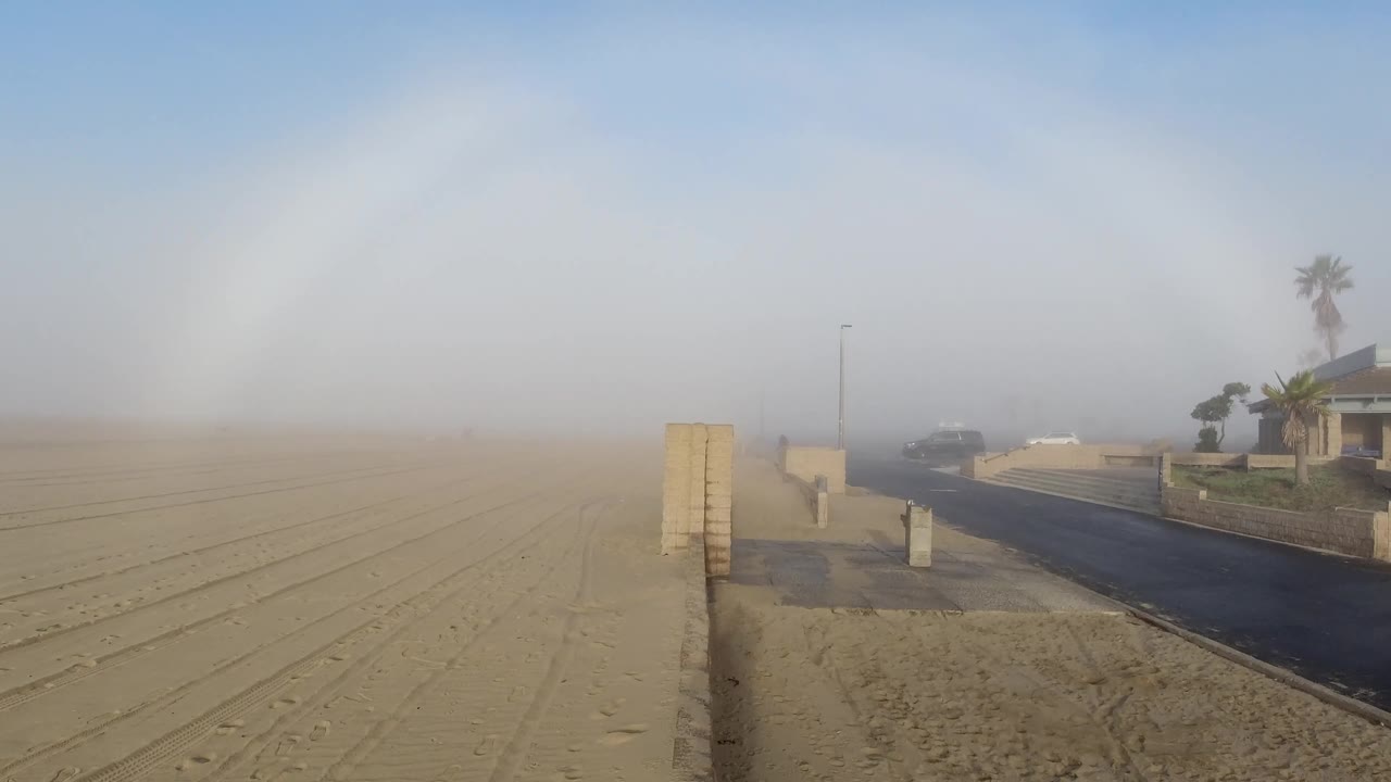 At 0:02 in Huntington Beach, open sand stretches toward a hazy horizon while a roadside wall, a palm-lined home, and parked vehicles sit along the right side, with no visible people.