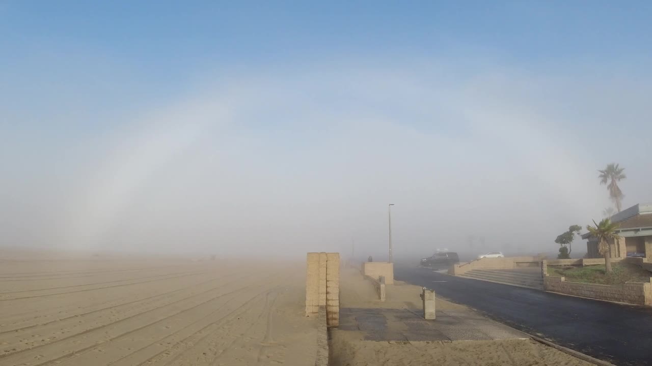 Opening frame at Huntington Beach shows a full white fogbow arc spanning the sky above a fog-laced residential beach street; a concrete bollard sits in the center foreground and a palm tree with low rooflines appears in the right midground, and no people are visible