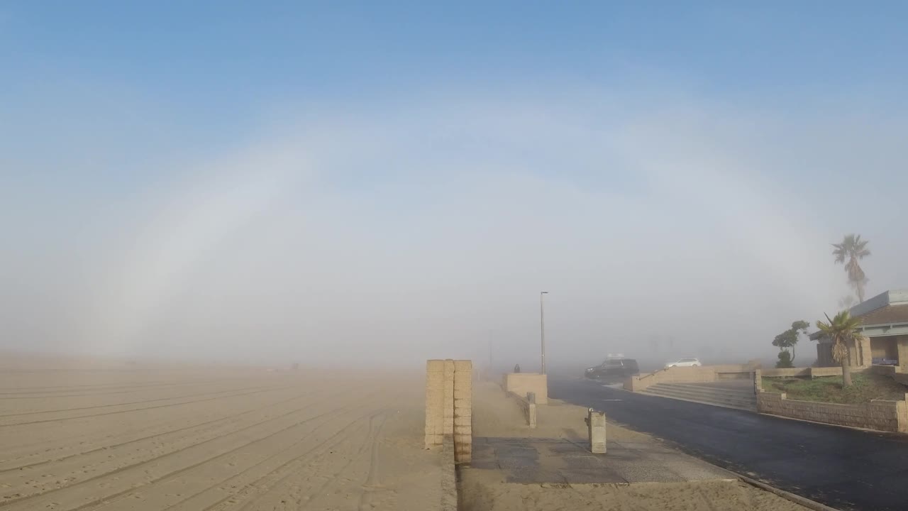 Two seconds in, the fogbow arc spans from upper left to upper right above the Huntington Beach residential street; the white beach house in the right midground is the most legible structure in the frame, a light pole stands near the bollard, and no people are visible
