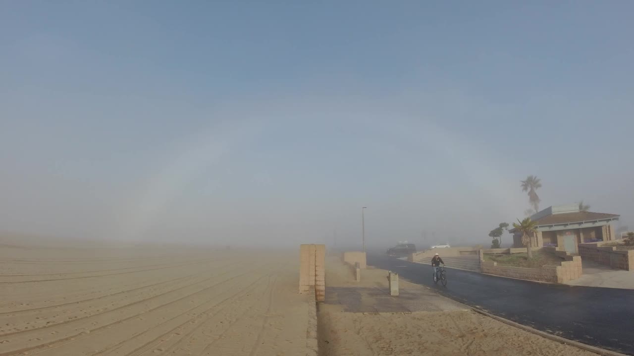 Cyclist on a dark road beneath a faint rainbow near a sandy area.