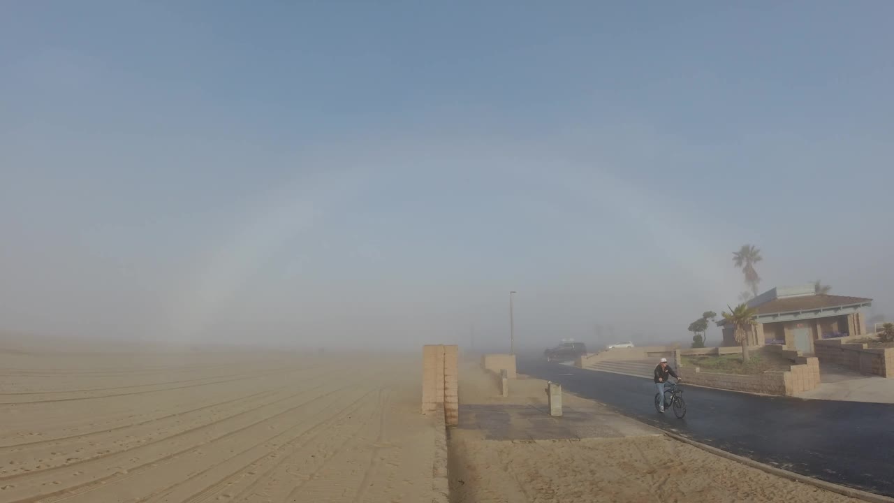 A cyclist rides along the right side of the road near a low wall and sandy open ground, with a house, palm trees, parked cars, and a pale rainbow in the misty Huntington Beach sky.