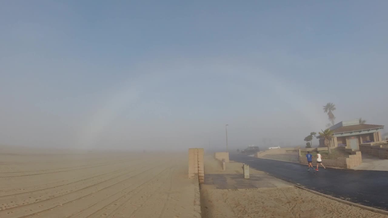 At Huntington Beach, two small people are visible near a paved path as windblown sand hazes the beach beside a house and palm trees.