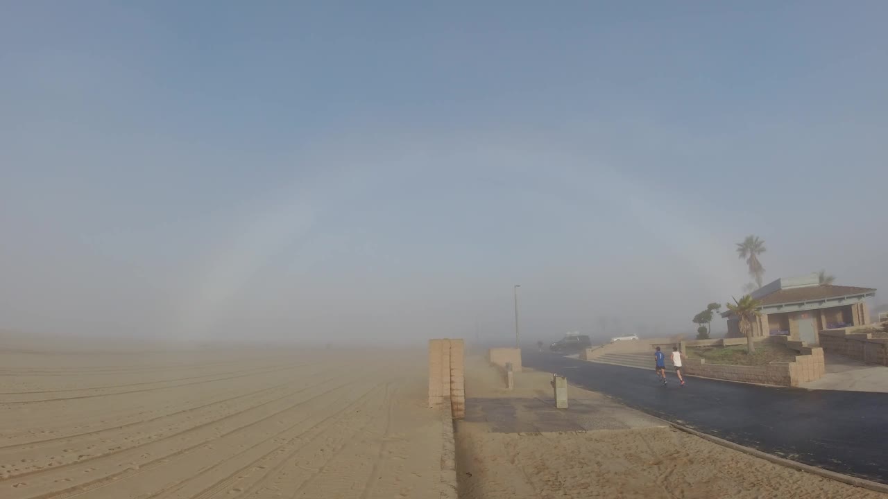 At Huntington Beach, two people are farther along the dark path near a beach house, with windblown sand softening the open shoreline to the left.