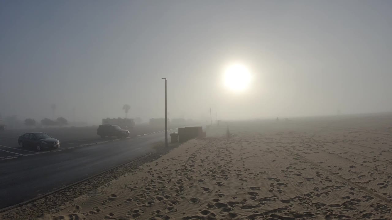 Opening frame of Huntington Beach looking across footprint-covered sand toward a sun diffused into a bright orb through dense marine layer haze, with dark parked cars along the road at the far left and a light pole nearby; no people visible.