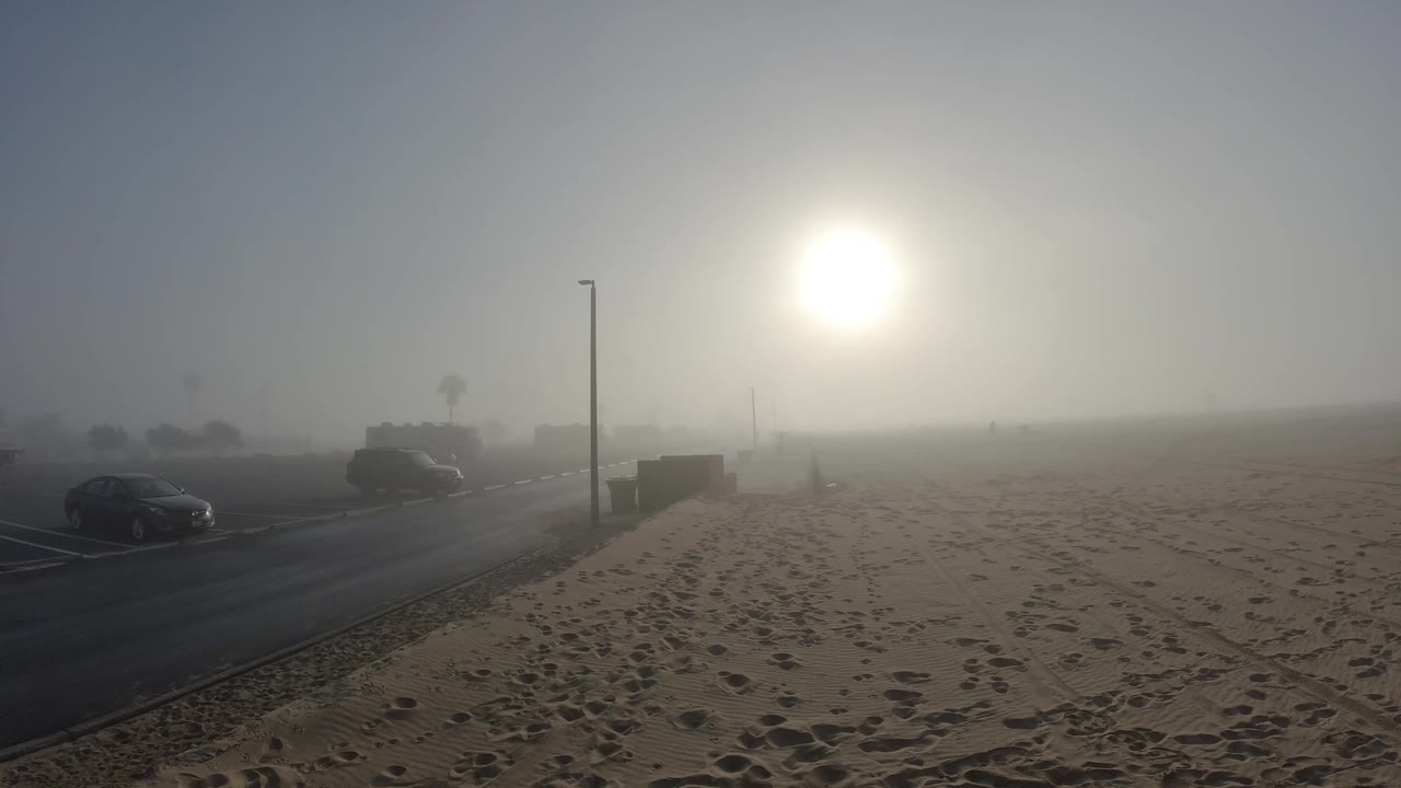 One second in, a dark parked car at the road edge is more distinctly framed than in the opening frame, the light pole stands left of center, and the sun glows upper-right through the marine layer at Huntington Beach; no people visible.