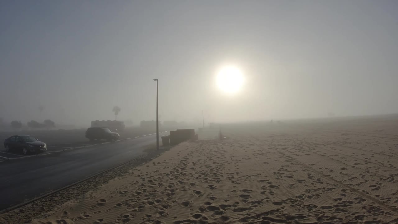 At two seconds, the Huntington Beach composition holds with the light pole standing just left of center and the sun maintaining its diffused glow; the road and parked cars at the left edge are static, and the footprint-covered foreground remains unchanged with no people visible.
