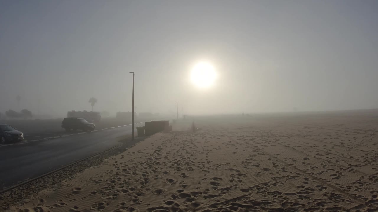 Sandy beach with footprints, road and parked cars at left, street light pole, hazy sun, and a second light pole faintly visible in background haze.