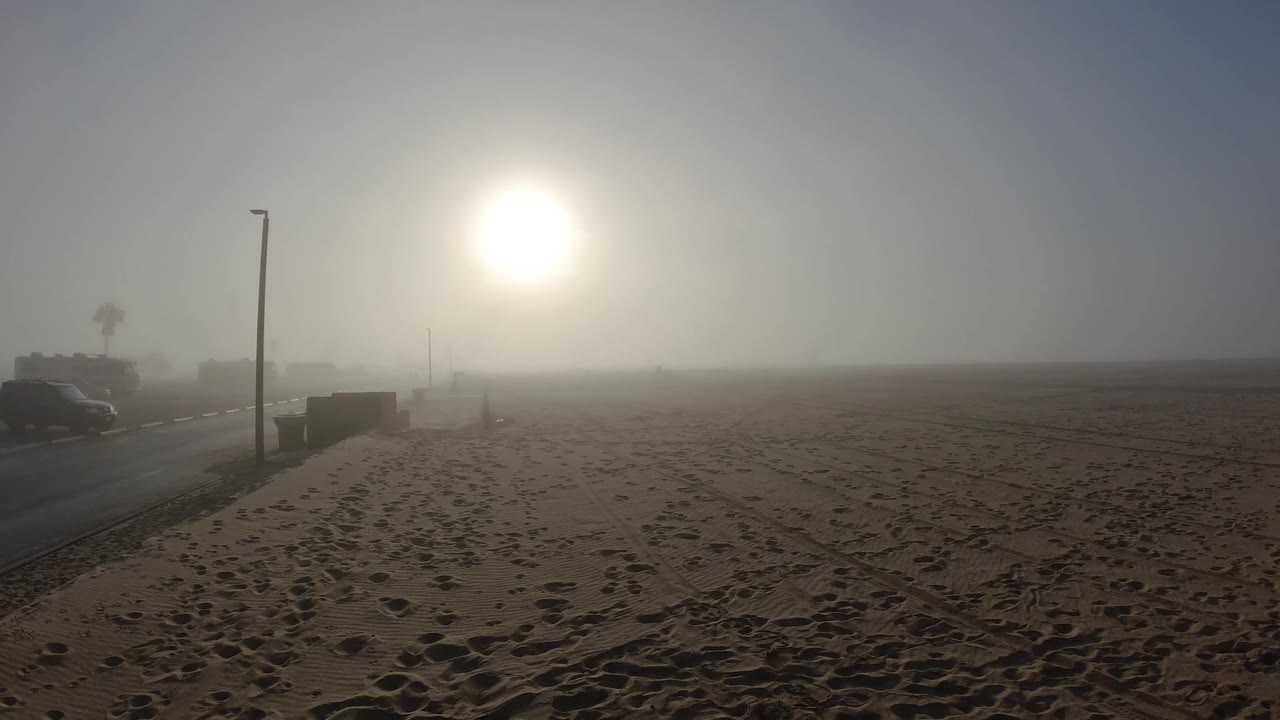 Opening frame at Huntington Beach, California: bright white sun burns through coastal haze in the upper center-left, a dark lamp post stands at frame left, footprint tracks crisscross the foreground sand, and a low concrete barrier sits in the midground; no people are visible.