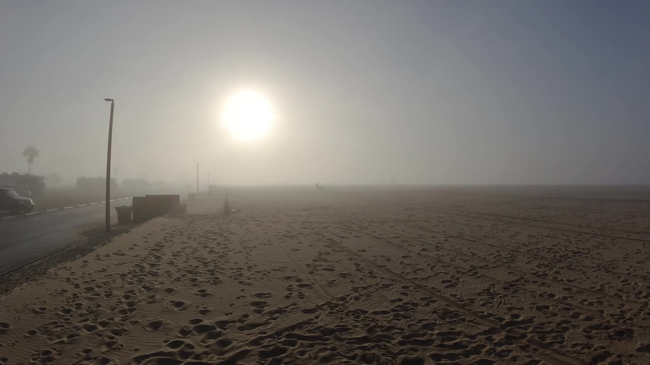 At 0:02 at Huntington Beach, dark vegetation silhouettes behind the lamp post are marginally more defined in the upper-left corner compared to the first two frames; the sun glare, foreground footprint tracks, and concrete barrier remain consistent; no people are visible.