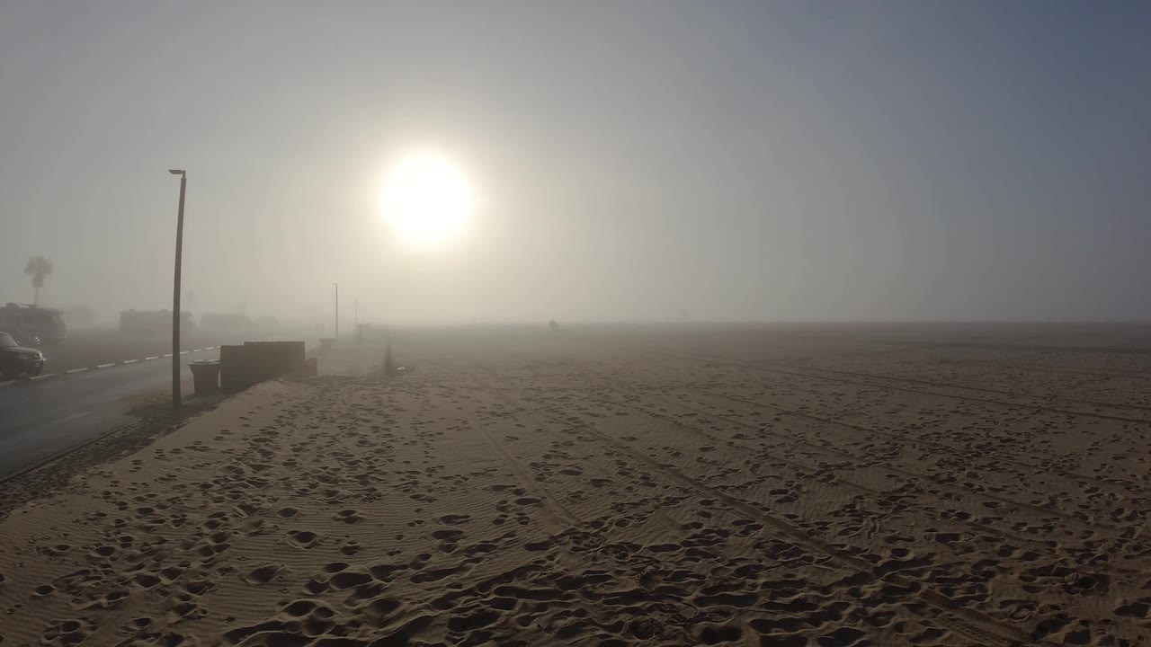 Beach midpoint frame with hazy sun, lamp post, and wide expanse of footprint-patterned sand; haze appears slightly denser near the horizon.