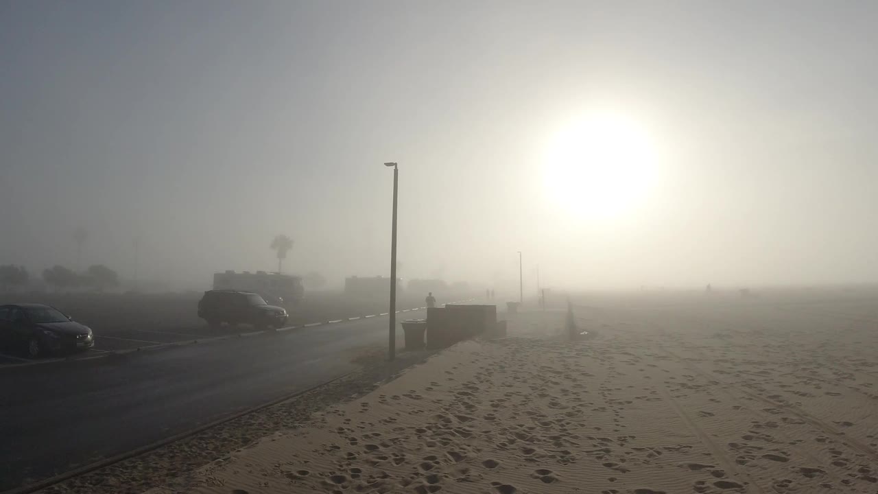 A wide Huntington Beach view shows footprints across the sand, a road with dark vehicles, tall poles, and a distant palm shape softened by fog.