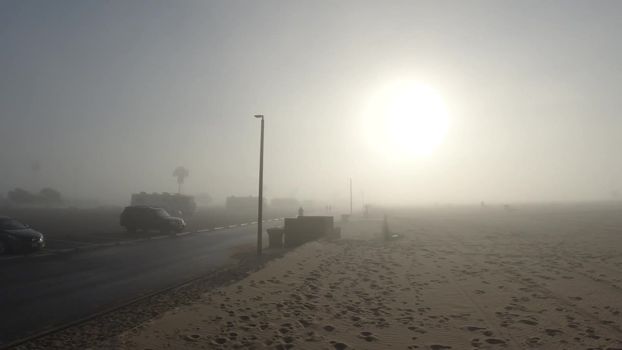 The Huntington Beach shoreline area is mostly empty sand with a dark vehicle near the road, a vertical light pole, distant buildings, and strong sun glare.