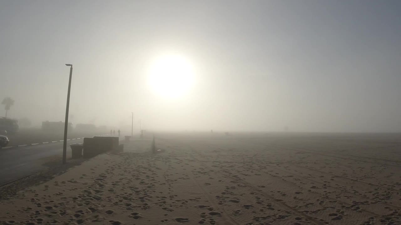At Huntington Beach, footprint-covered sand fills the foreground, a tall lamp post stands at the left edge, and the sun appears as a bright diffused orb through dense coastal fog at the clip's opening; no people are visible