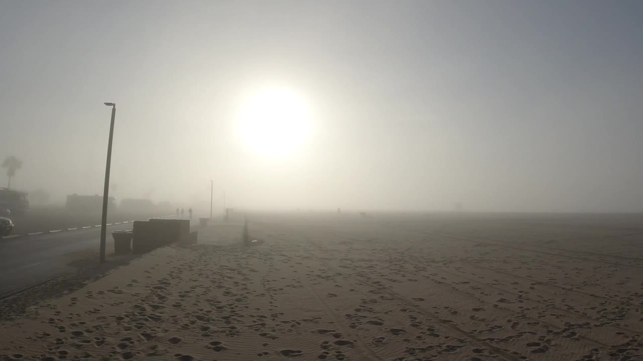 One second into the Huntington Beach clip, the footprinted sand stretches to the fog-obscured horizon, the lamp post remains at the left, and the diffused sun sits slightly more centered in the frame than in the opening; no people are visible