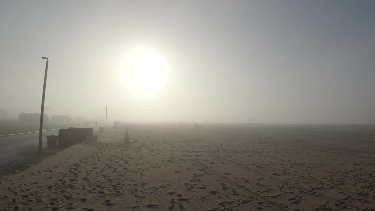 Two seconds in at Huntington Beach, the lamp post has shifted toward the center of the frame as the camera moves, the sun glows in the upper right area, and the footprinted sand and concrete path edge remain consistent across the foreground and midground; no people are visible