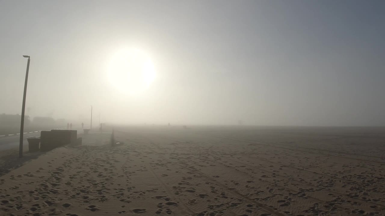 Sandy beach, lamp post at left, sun through noticeably thicker fog, concrete barrier more prominent at lower left
