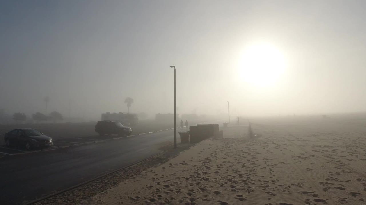 Opening frame of a foggy Huntington Beach morning: sandy beach with footprints fills the foreground, a parked dark car sits left of a metal street lamp in the midground, and a bright sun glows as a diffuse white orb through dense marine fog; no people visible.