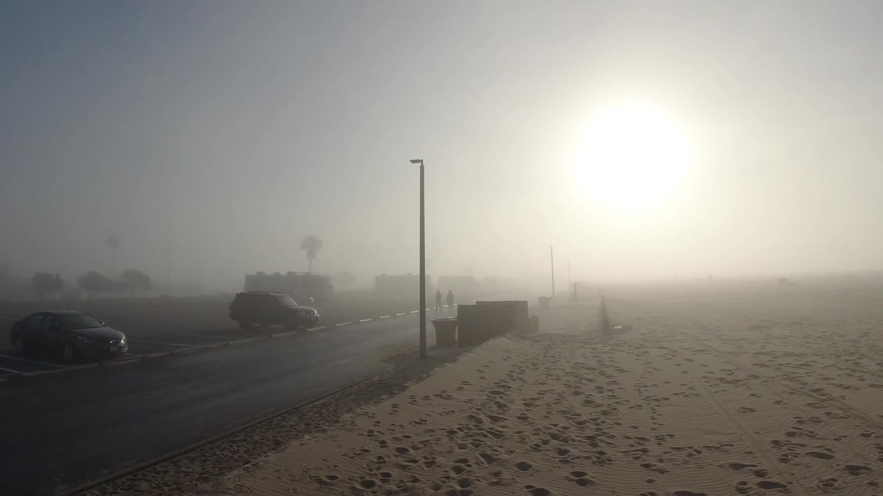 Foggy beach road with dark parked car and street lamp, sun glowing through marine fog over sand