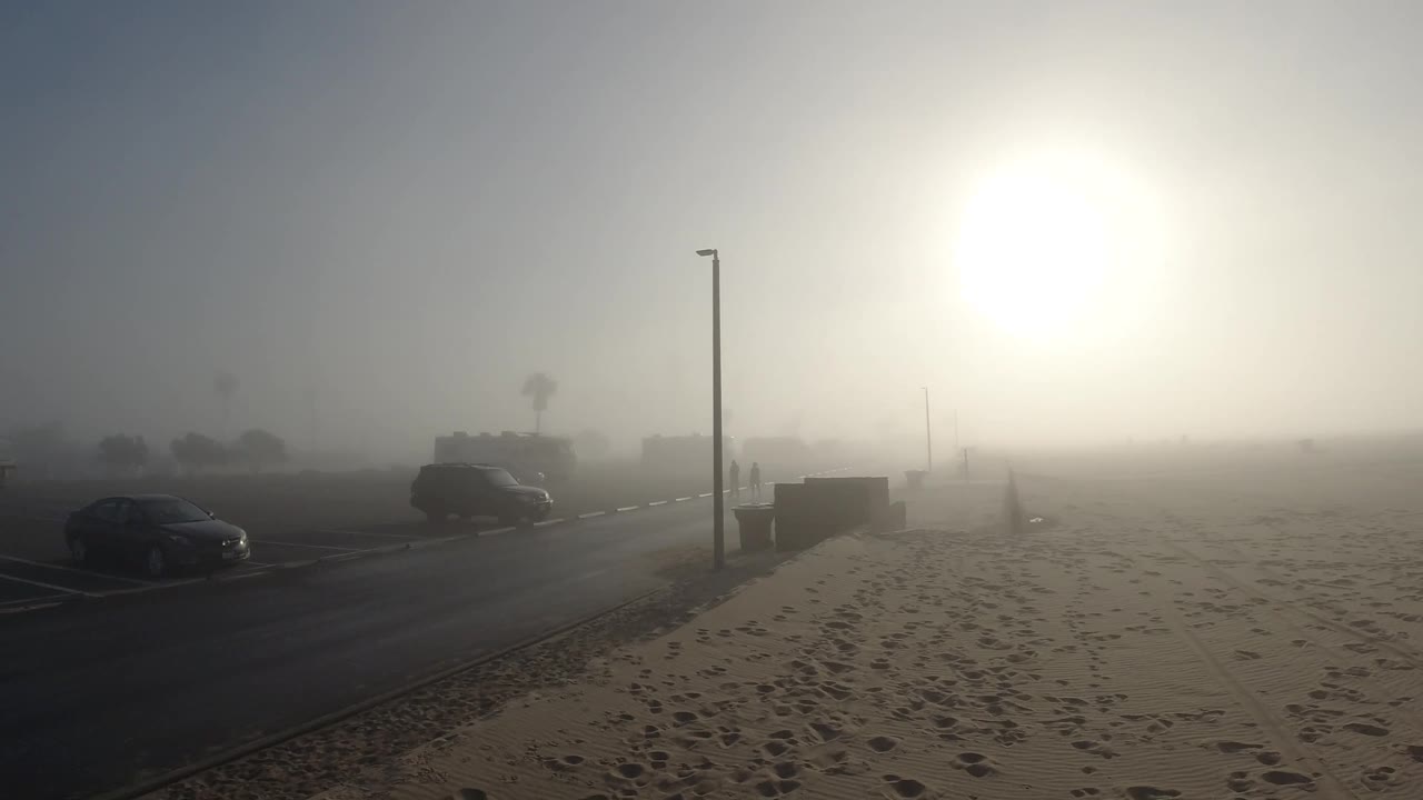 At two seconds into the Huntington Beach clip, the diagonal road cuts through the frame with the street lamp prominent center-right, the parked car in the midground, and the sun glowing as a diffuse white orb through the marine fog while the sandy foreground shows footprints; no people visible.
