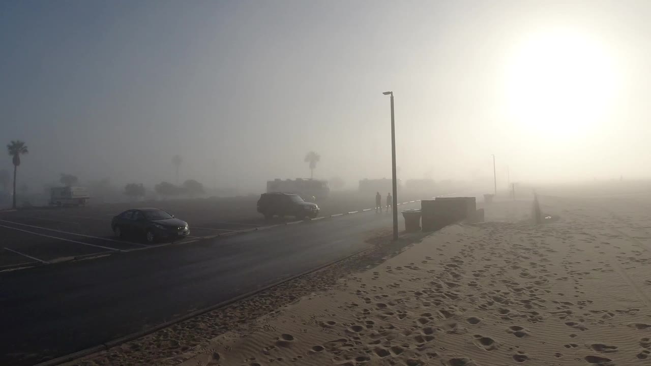 Huntington Beach, California beachside street seen across sandy foreground, with parked cars, palm trees, bright sun haze, and no visible people in the opening frame.